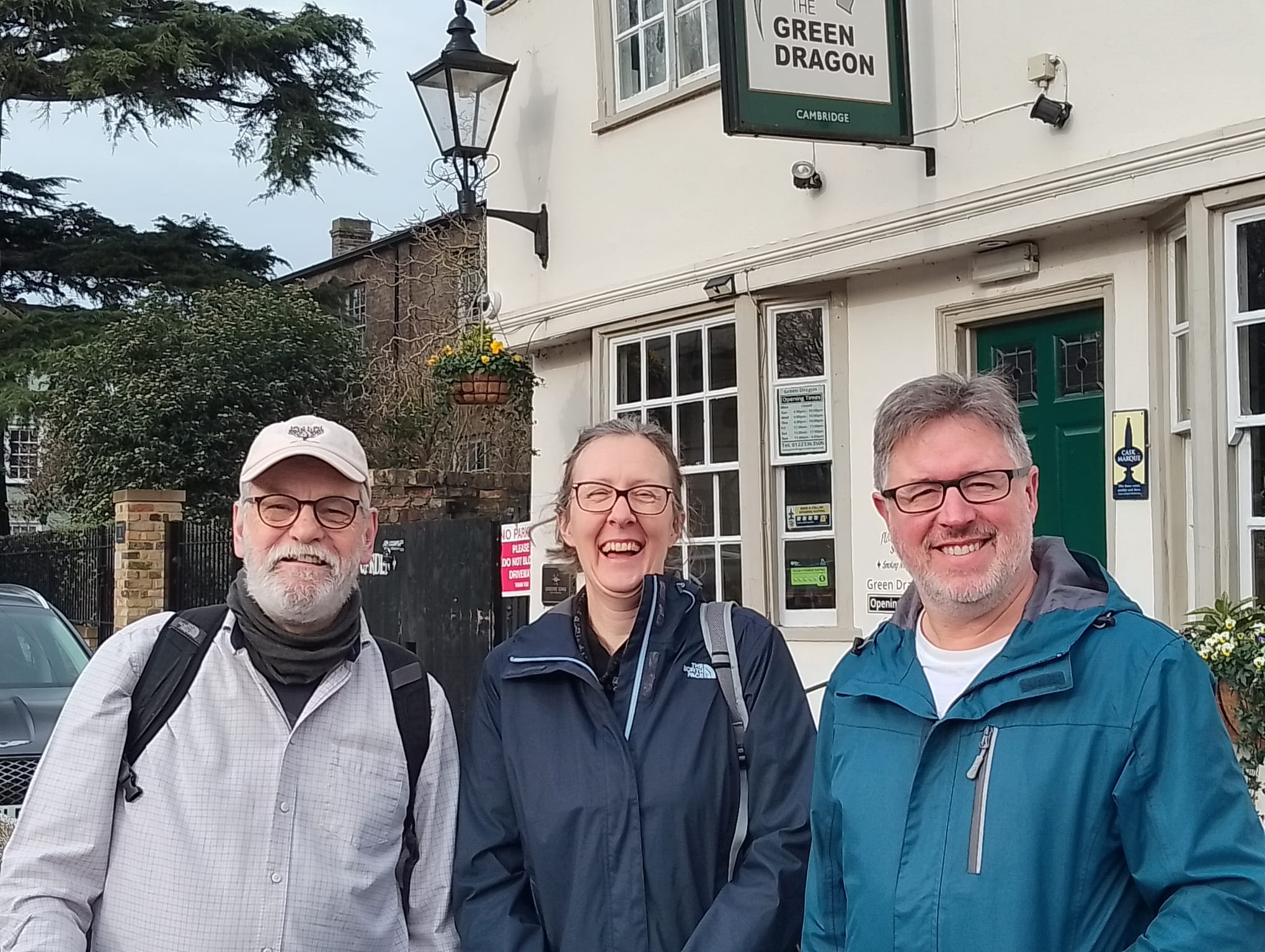 Two men and one woman stood outside of a cream pub with a green trim.
